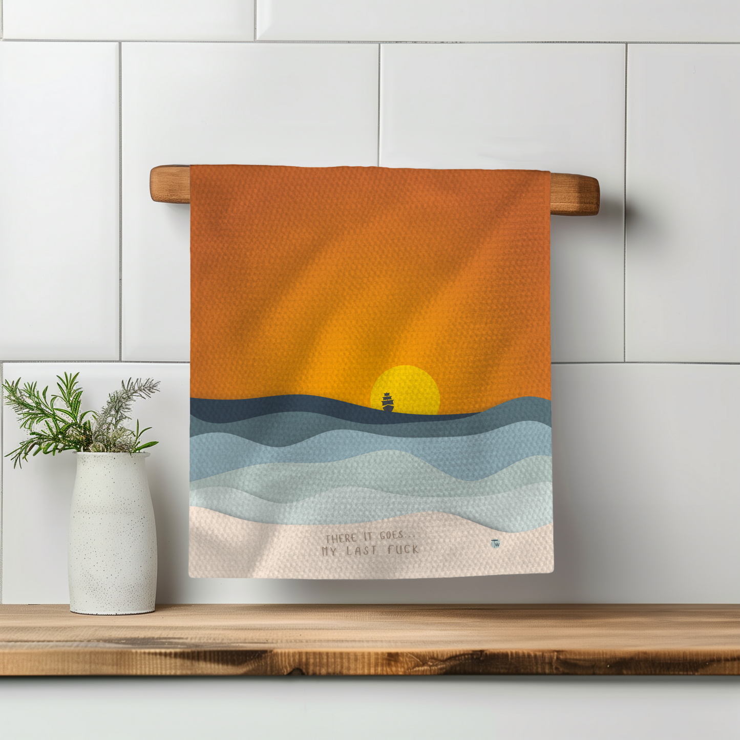 There It Goes Perfect Towel with an orange sunset and waves design, hanging on a wooden rack in a kitchen setting.