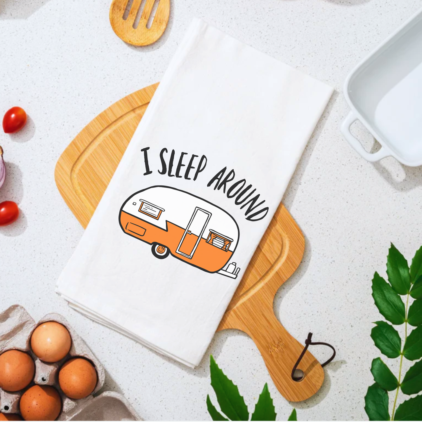 I Sleep Around tea towel featuring an orange camper illustration on a white background, placed on a wooden cutting board.