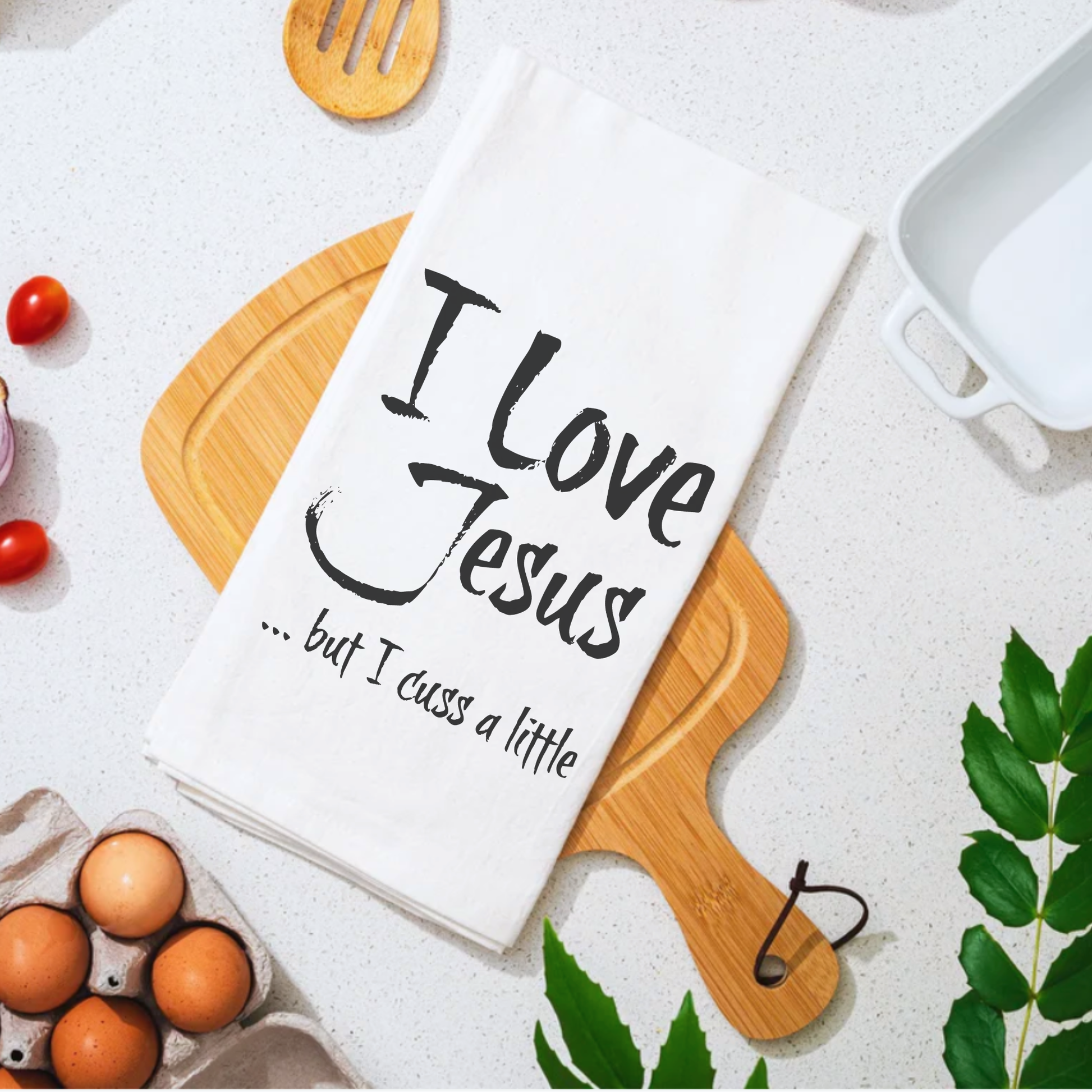 White cotton tea towel with the text "I Love Jesus ... but I cuss a little" displayed on a wooden cutting board.