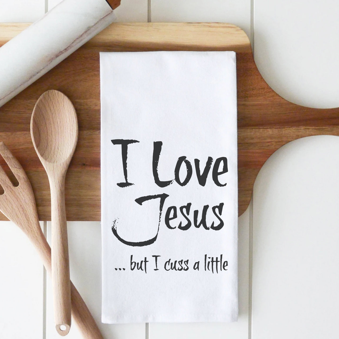 White cotton tea towel with black text reading "I Love Jesus but I cuss a little," displayed on a wooden cutting board.
