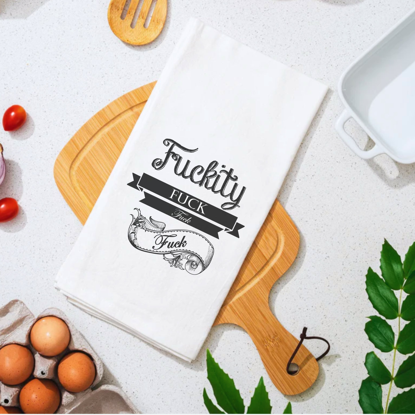 White cotton tea towel featuring the text "Fuckity" and decorative elements, placed on a wooden cutting board with food it...