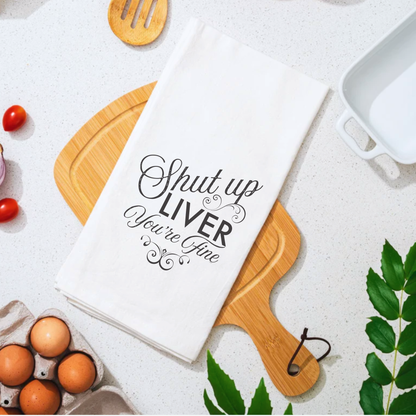 Shut up liver you're fine cotton tea towel on a wooden cutting board with eggs and tomatoes in the background.