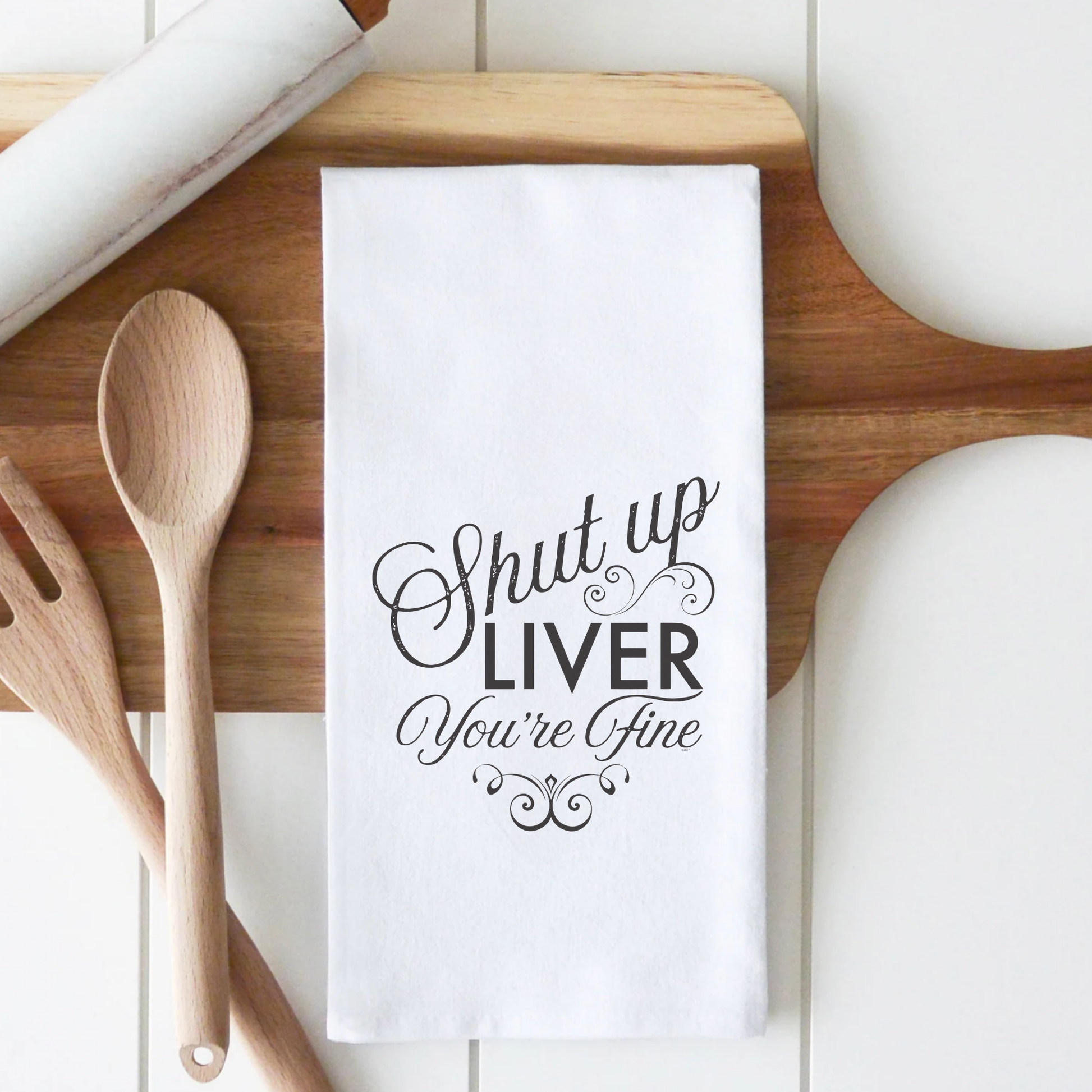 White cotton tea towel with the text "Shut up LIVER You're Fine" in black decorative font, placed on a wooden surface.