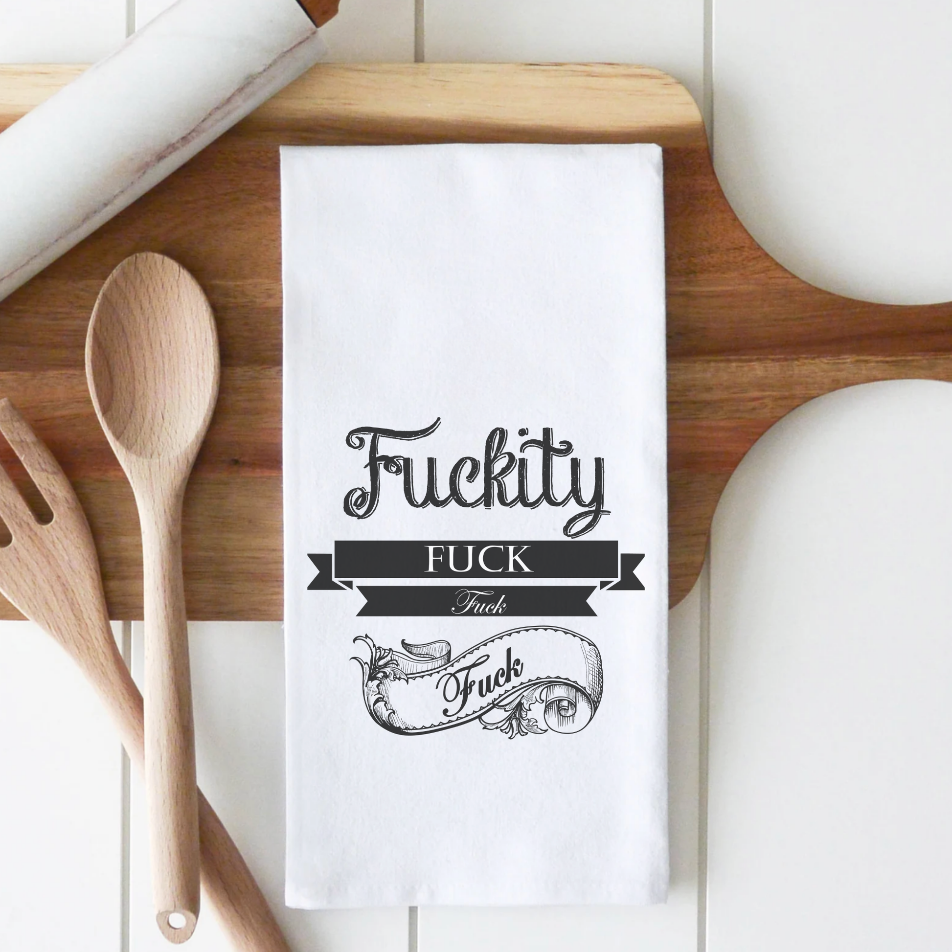 White cotton tea towel featuring the text "Fuckity Fuck" in decorative black font, placed on a wooden surface with utensils.