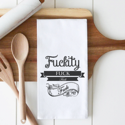 White cotton tea towel featuring the text "Fuckity Fuck" in decorative black font, placed on a wooden surface with utensils.
