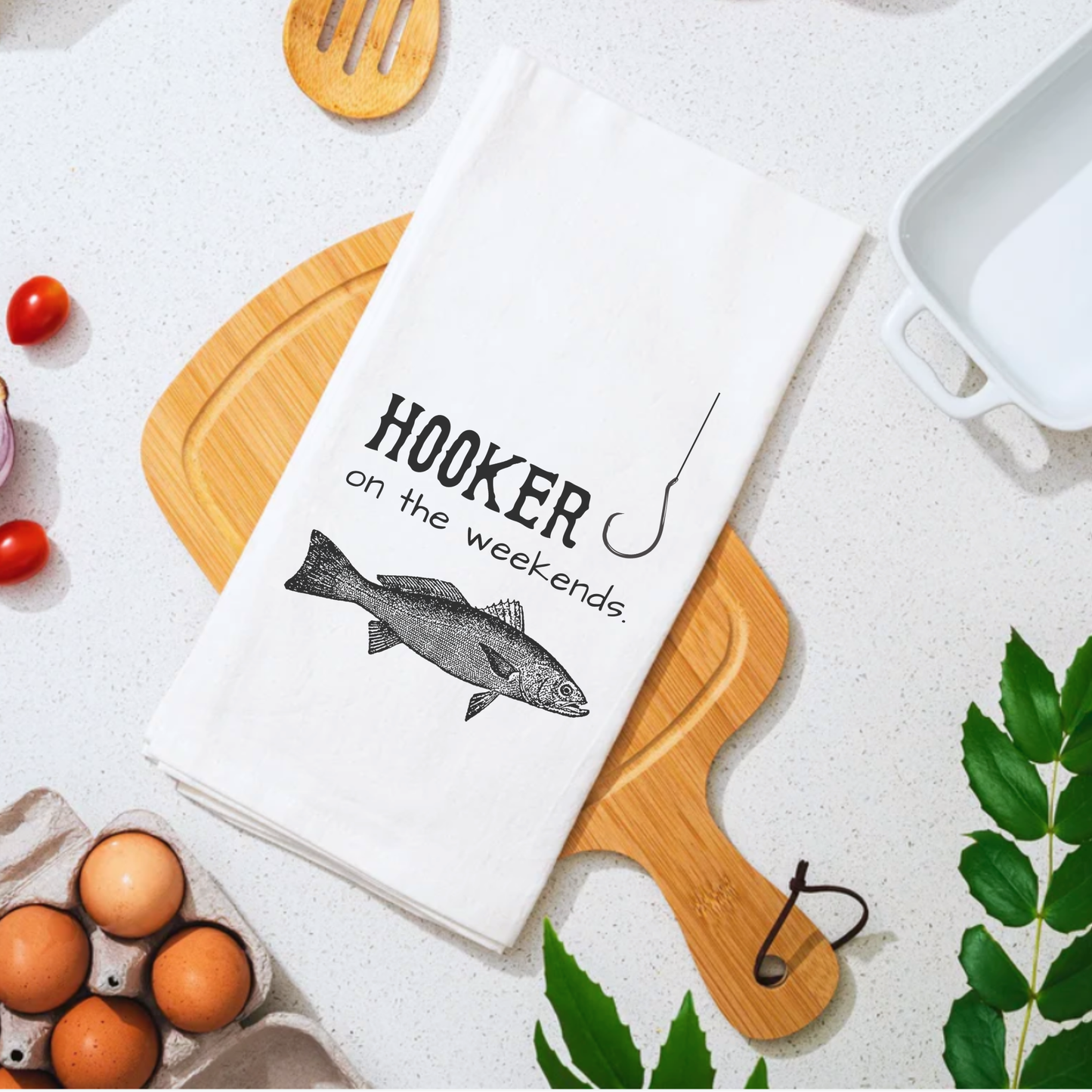 White cotton tea towel featuring the text "Hooker on the weekends" and an illustration of a fish, placed on a wooden cutti...