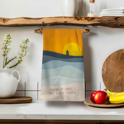 There It Goes Perfect Towel in orange and blue, featuring a sunset and ship design, hanging on a wooden shelf.