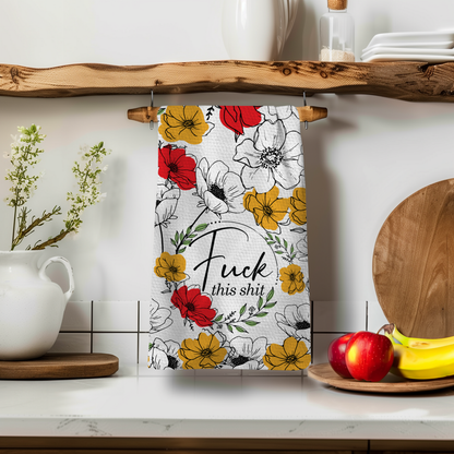 Floral towel featuring the phrase "Fuck this shit" in black, hanging on a wooden shelf with kitchen decor.