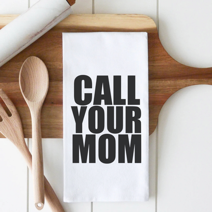 White cotton tea towel with bold black text saying "CALL YOUR MOM," placed on a wooden cutting board with utensils.