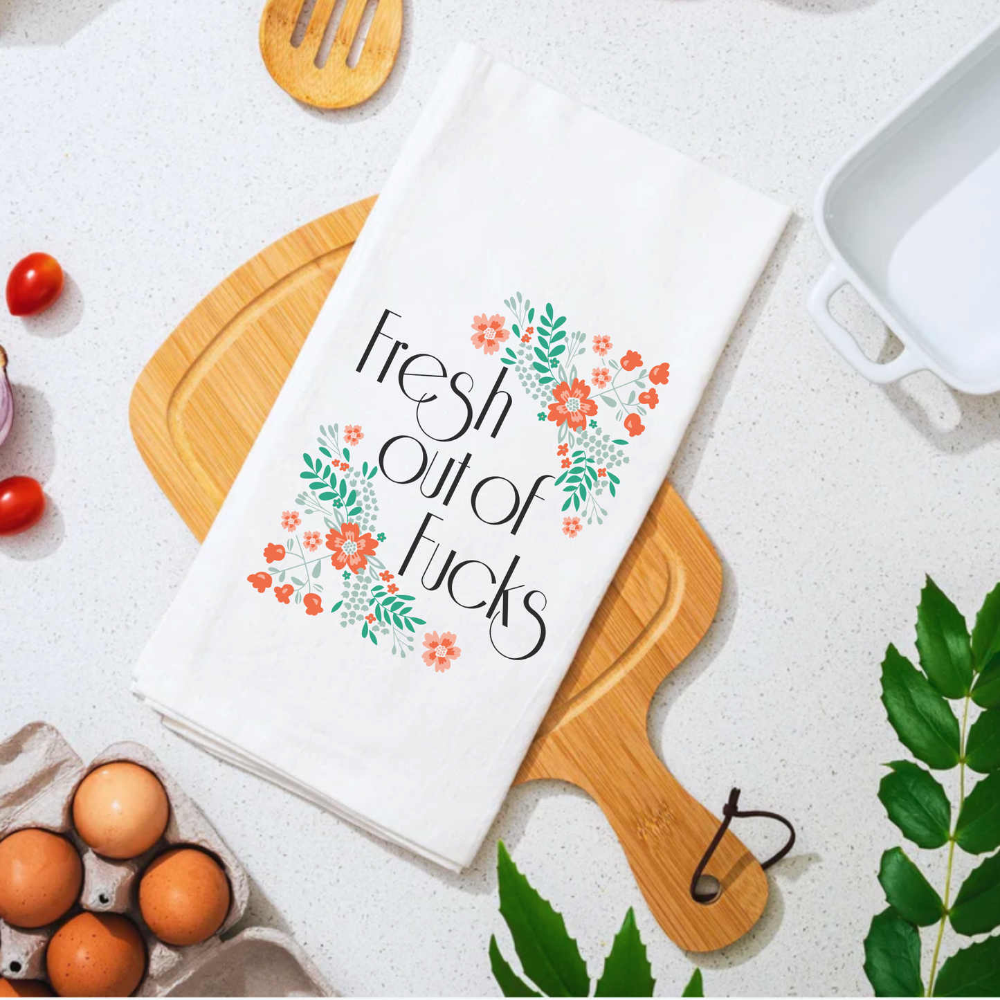 White cotton tea towel with "Fresh Out of Fucks" text and floral design, placed on a wooden cutting board.