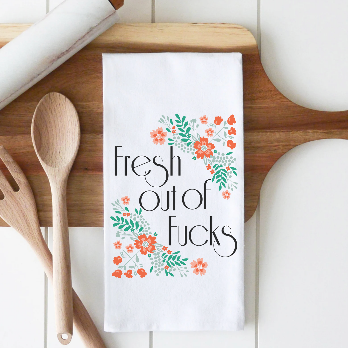 Fresh Out cotton tea towel featuring floral design and bold text on a wooden countertop with kitchen utensils.