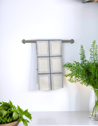 Fresh Out Plaid Illusion kitchen tea towel in cream and gray hanging on a metal rack, with greenery in the background.