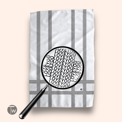 White towel featuring a striped design with repeated text "Love You" in black, magnified under a black magnifying glass.
