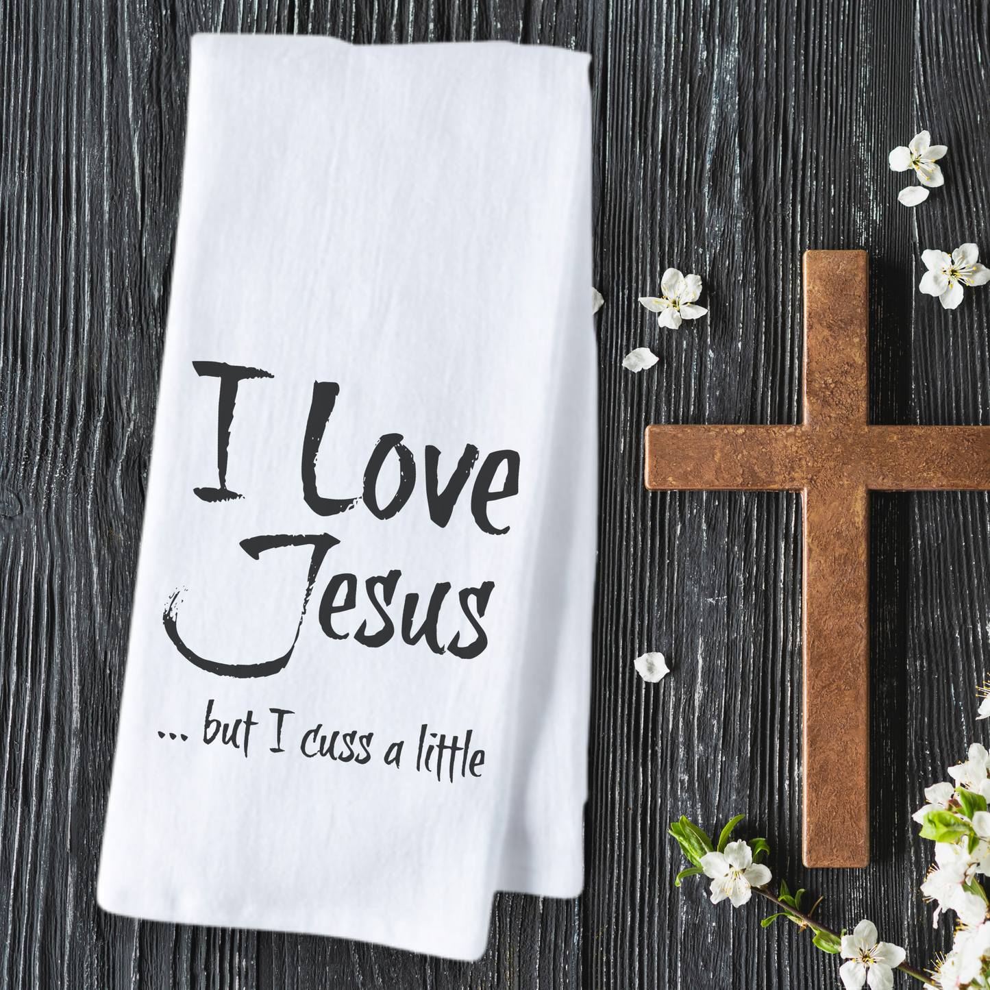 White cotton tea towel featuring the text "I Love Jesus but I cuss a little" on a wooden background with a cross and flowers.