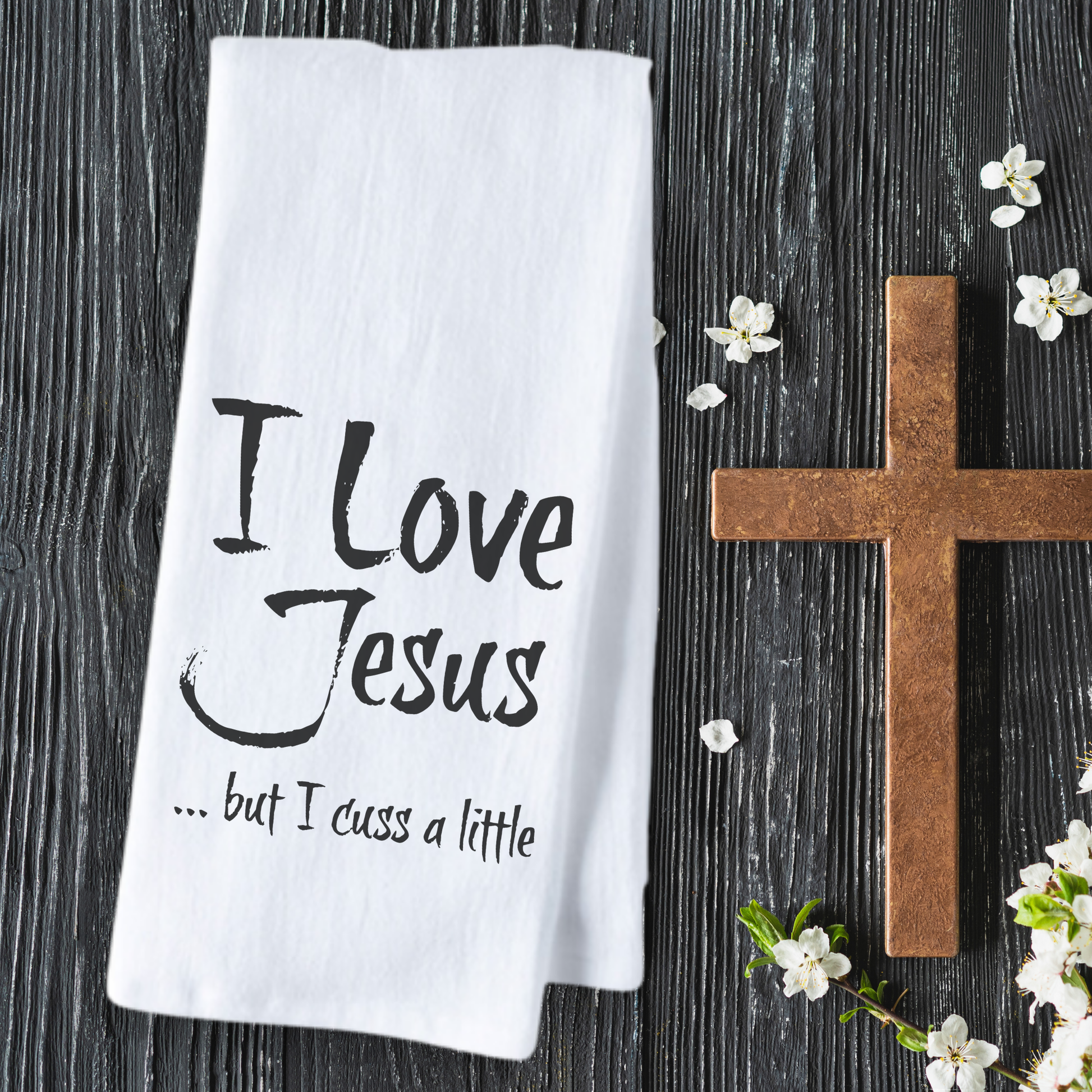 White cotton tea towel featuring the text "I Love Jesus but I cuss a little" on a wooden background with a cross and flowers.