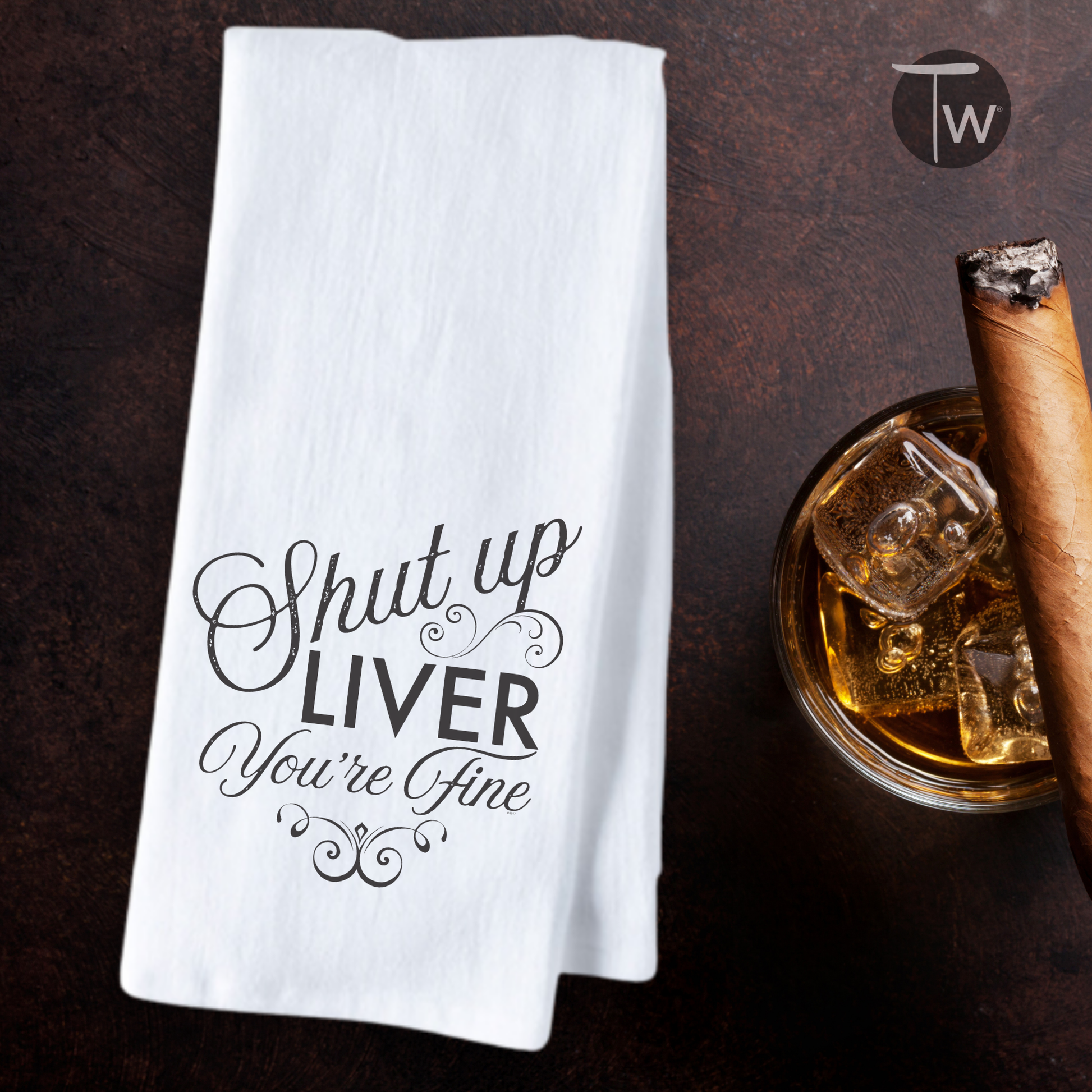 White cotton tea towel featuring the phrase "Shut up Liver You're Fine" in decorative black lettering, placed beside a gla...