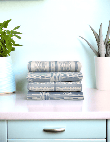 Fresh Out Plaid Illusion kitchen tea towels stacked in shades of blue and gray on a countertop with plants in the background.